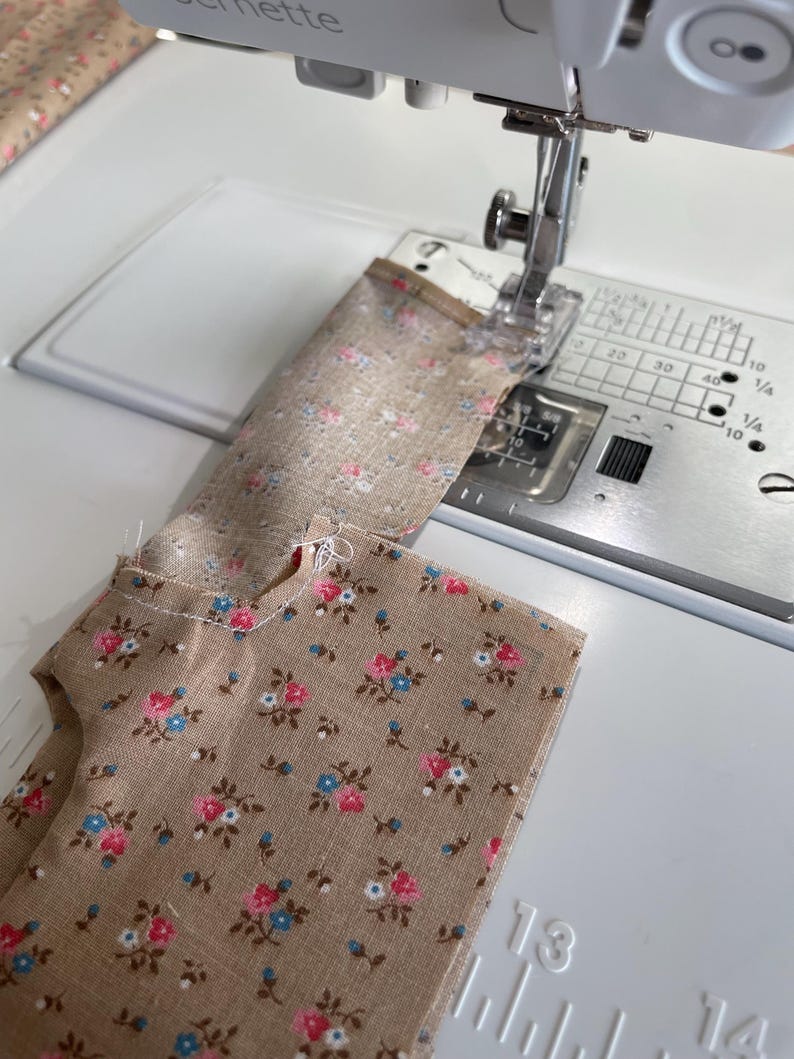 May include: A sewing machine stitching floral fabric. The fabric is tan with a pattern of pink, blue, and white flowers. The machine is white with silver components. The fabric is being sewn in a straight line.