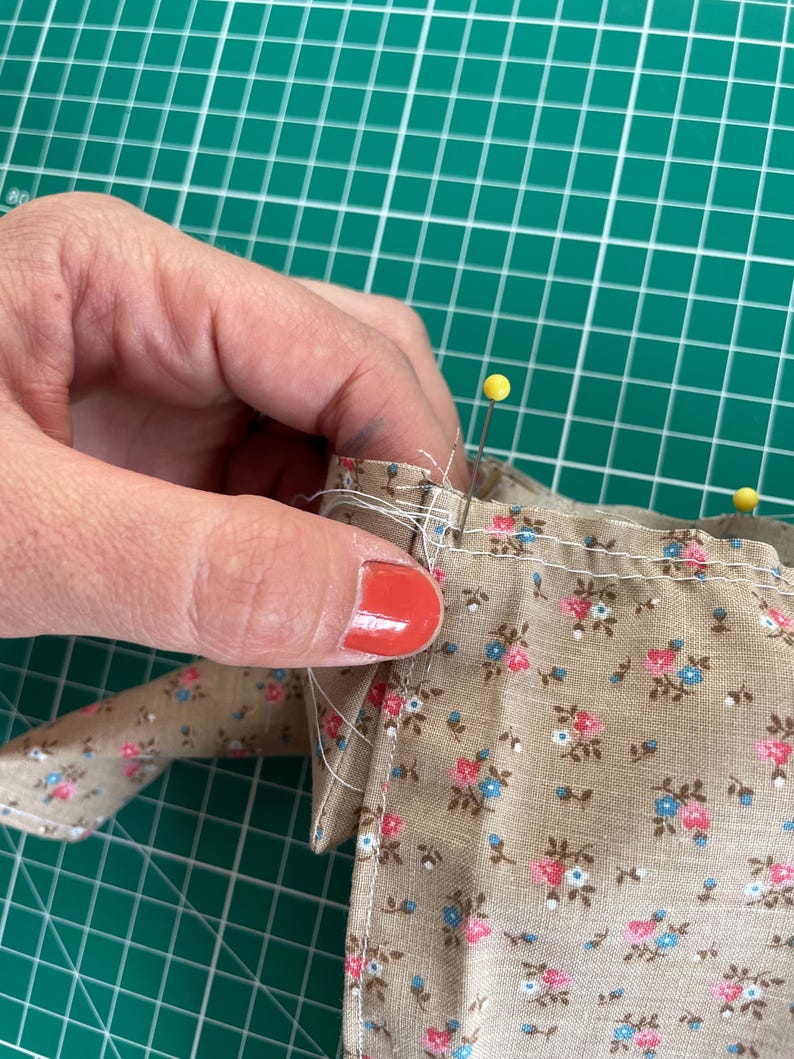 May include: Close-up of fabric with a floral pattern in pink and blue on a tan background. Yellow pins secure the fabric edges. The fabric is being sewn, showcasing a sewing project.