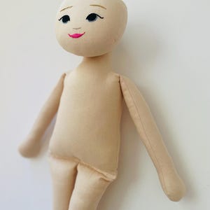 May include: A handmade cloth doll with a beige fabric body and limbs. The doll's face is embroidered with black eyes, eyebrows, and a pink mouth. The doll's arms are outstretched, and it stands against a white background.