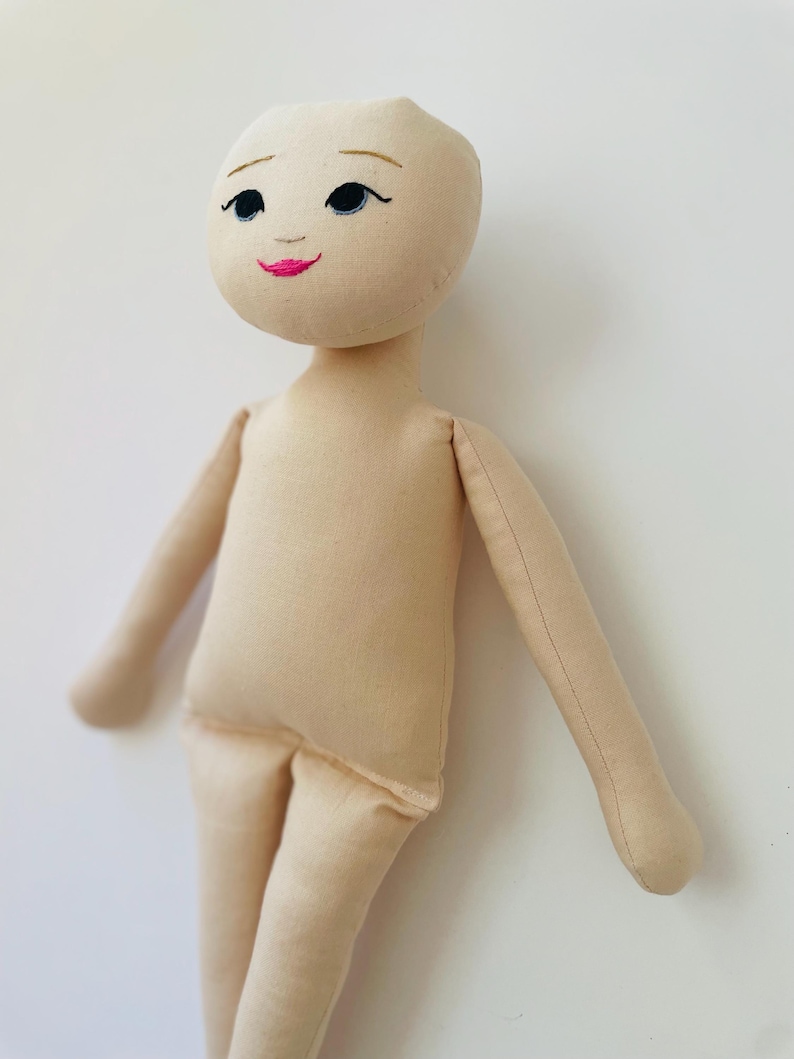 May include: A handmade cloth doll with a beige fabric body and limbs. The doll's face is embroidered with black eyes, eyebrows, and a pink mouth. The doll's arms are outstretched, and it stands against a white background.