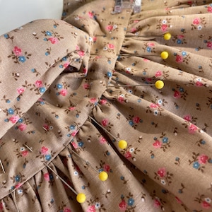 May include: Close-up of a sewing project featuring a tan fabric with a floral pattern in pink and blue. Yellow pins secure the fabric, which is being sewn by a sewing machine. The fabric is gathered and appears to be part of a garment.