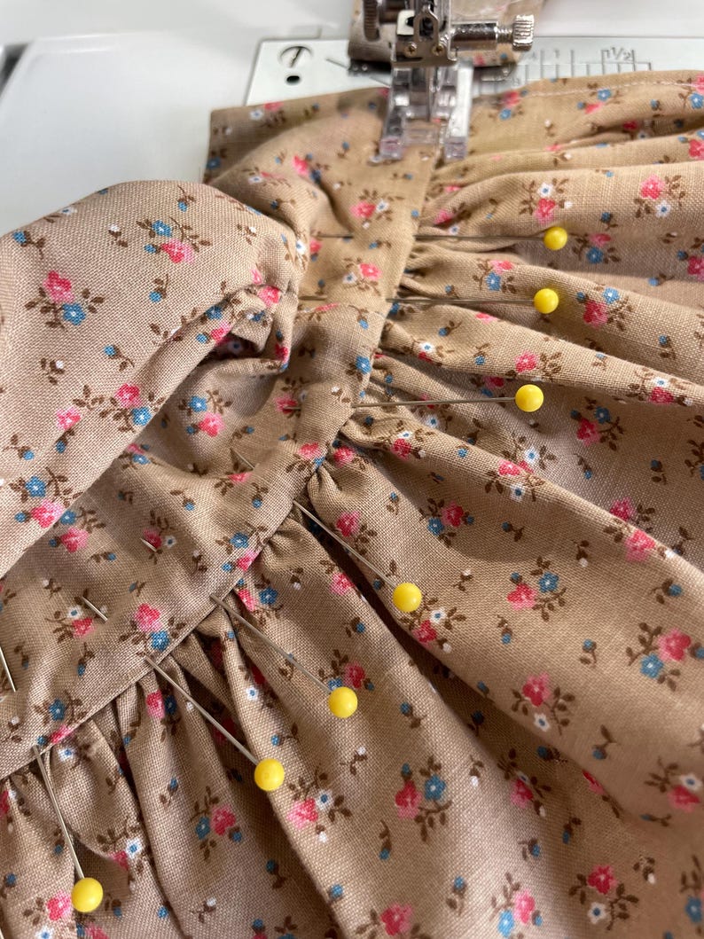 May include: Close-up of a sewing project featuring a tan fabric with a floral pattern in pink and blue. Yellow pins secure the fabric, which is being sewn by a sewing machine. The fabric is gathered and appears to be part of a garment.