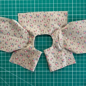May include: Fabric pieces for a garment, featuring a floral pattern in pink, blue, and green on a tan background. The fabric is cut into sections for the neckline and sleeves, ready for sewing. The pieces are laid out on a green cutting mat.