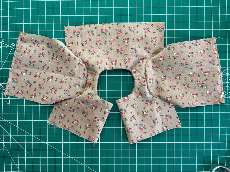 May include: Fabric pieces for a garment, featuring a floral pattern in pink, blue, and green on a tan background. The fabric is cut into sections for the neckline and sleeves, ready for sewing. The pieces are laid out on a green cutting mat.
