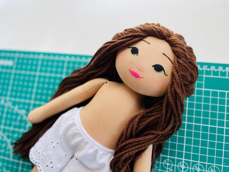 May include: A handmade doll with brown yarn hair and a fabric face. The doll has a light skin tone, black eyes, and a pink mouth. It is wearing white shorts with eyelet detailing. The doll is lying on a green cutting mat.