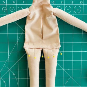 May include: A beige fabric doll body with arms and legs pinned with yellow-headed pins. The doll body is on a green cutting mat with measurements in inches.