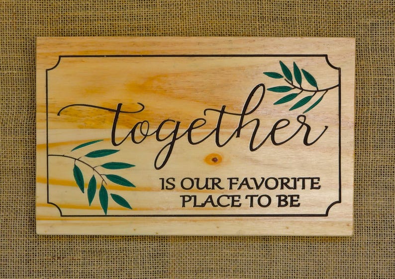 May include: Wooden sign with the text "Together is our favorite place to be" in black lettering. The sign has a light wood background and green leafy branches on either side of the text.