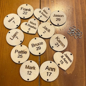 May include: A collection of wooden tags in various shapes, including circles and hearts. Each tag is engraved with a name and a number. Some tags have names like "Frank" and "Mary". Small metal hooks are also present.