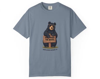 May include: A blue short-sleeve t-shirt featuring a cartoon bear wearing sunglasses and holding a sign that reads "DO NOT FEED THE BEARS." The shirt also includes the text "BARK EATER OUTDOOR CO."