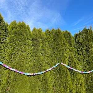 Pu&ograve; includere: Una ghirlanda di perline colorate che compone la parola "TINSEL" &egrave; appesa su uno sfondo di cespugli sempreverdi verdi.