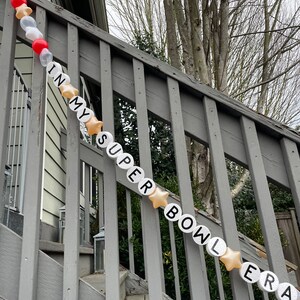 May include: A white and gold balloon banner spelling out "IN MY SUPER BOWL ERA" hangs on a wooden railing. The banner is made of individual balloons with each letter printed on a separate balloon.