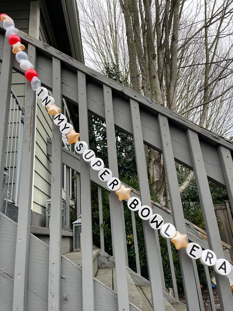 May include: A white and gold balloon banner spelling out "IN MY SUPER BOWL ERA" hangs on a wooden railing. The banner is made of individual balloons with each letter printed on a separate balloon.