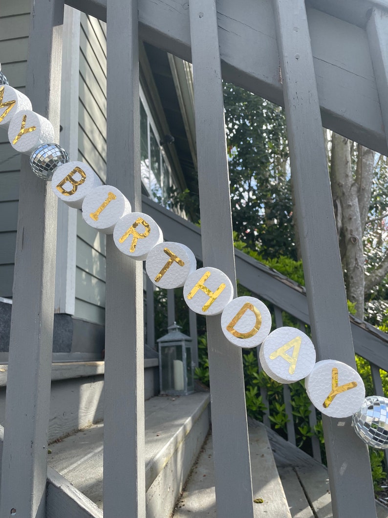 Puede incluir: C&iacute;rculos de espuma blanca con letras doradas que deletrean "Feliz cumplea&ntilde;os" est&aacute;n enhebrados con bolas de discoteca plateadas en una barandilla de madera gris.