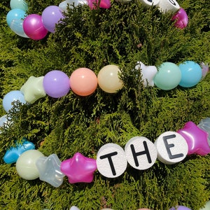 Pu&ograve; includere: Una ghirlanda di palline di plastica pastello e stelle bianche con le parole "TIS THE" scritte in lettere nere su cerchi bianchi.