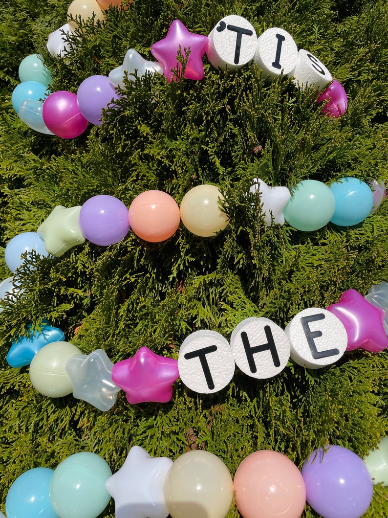 Pu&ograve; includere: Una ghirlanda di palline di plastica pastello e stelle bianche con le parole "TIS THE" scritte in lettere nere su cerchi bianchi.