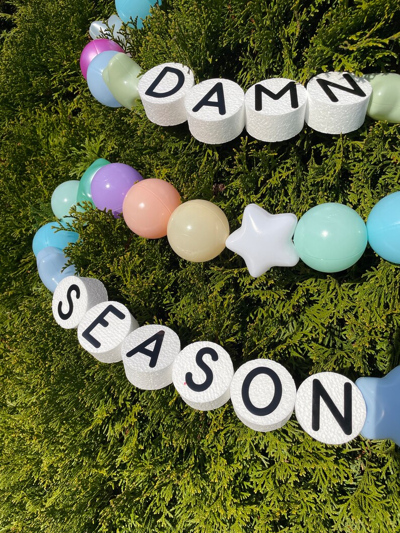 Pu&ograve; includere: Una ghirlanda di palloncini color pastello con lettere in schiuma bianca che compongono la scritta "DAMN SEASON".