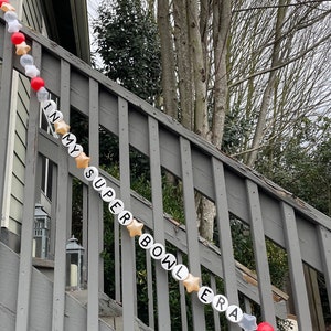 May include: A white and gold garland with the words "In My Super Bowl Era" strung on a wooden railing. The garland is decorated with red and white stars.