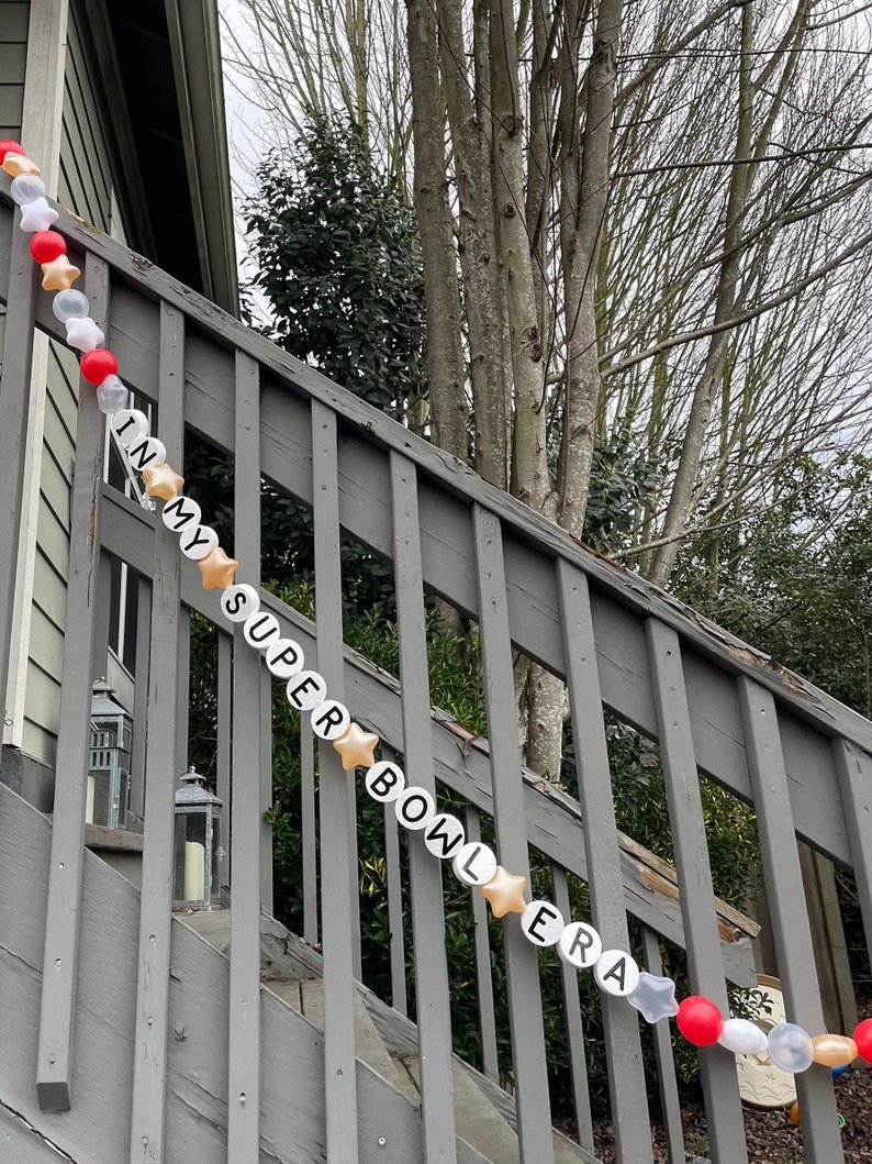 May include: A white and gold garland with the words "In My Super Bowl Era" strung on a wooden railing. The garland is decorated with red and white stars.