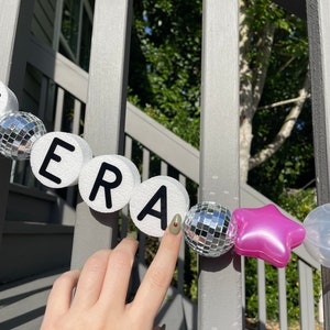 Puede incluir: Una pancarta de letras de espuma blanca que deletrea "ERA" con adornos de bola de discoteca plateada y un adorno de estrella rosa.