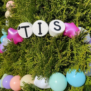 Pu&ograve; includere: Una ghirlanda bianca con la parola "'TIS" scritta in lettere nere su cerchi bianchi. La ghirlanda &egrave; decorata con stelle rosa e bianche e palloncini pastello.