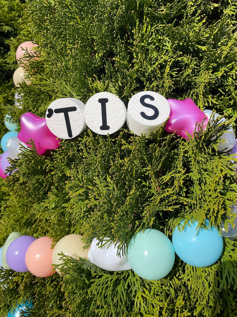 Pu&ograve; includere: Una ghirlanda bianca con la parola "'TIS" scritta in lettere nere su cerchi bianchi. La ghirlanda &egrave; decorata con stelle rosa e bianche e palloncini pastello.