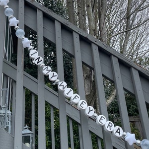 Może przedstawiać: Biały sznur koralik&oacute;w z czarnymi literami tworzącymi napis "In My Wife's Era" wiszący na szarej drewnianej balustradzie. Koraliki są przeplatane białymi gwiazdkami i przezroczystymi koralikami.