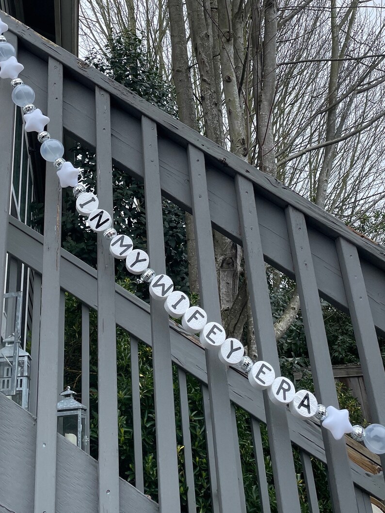 Może przedstawiać: Biały sznur koralik&oacute;w z czarnymi literami tworzącymi napis "In My Wife's Era" wiszący na szarej drewnianej balustradzie. Koraliki są przeplatane białymi gwiazdkami i przezroczystymi koralikami.
