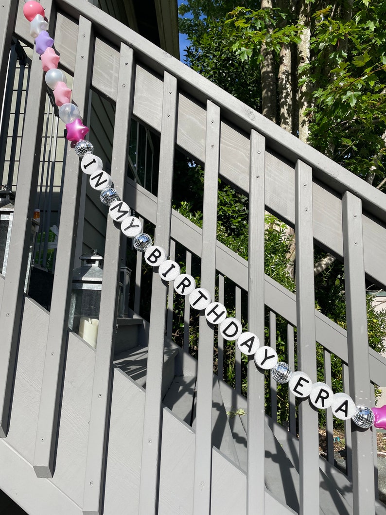 Puede incluir: Una pancarta de cumplea&ntilde;os plateada y rosa con las palabras "In My Birthday Era" en una escalera de madera.