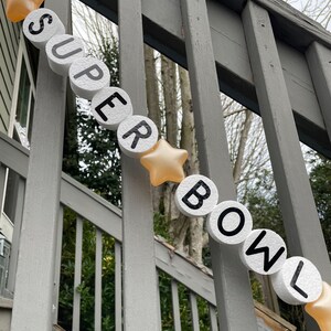 May include: A white foam banner with black letters spelling "SUPER BOWL" with gold stars between each letter. The banner is hanging on a gray wooden railing.