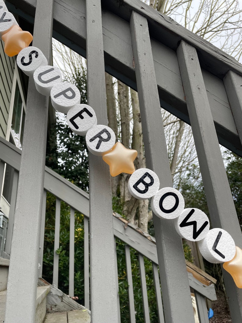 May include: A white foam banner with black letters spelling "SUPER BOWL" with gold stars between each letter. The banner is hanging on a gray wooden railing.
