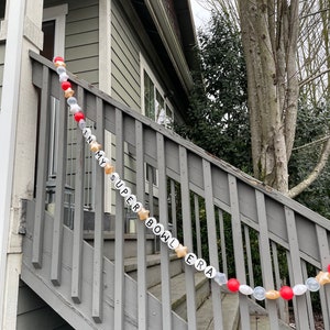 May include: A white and gold garland with the words "In My Super Bowl Era" strung on a wooden railing. The garland is decorated with red and gold balloons.