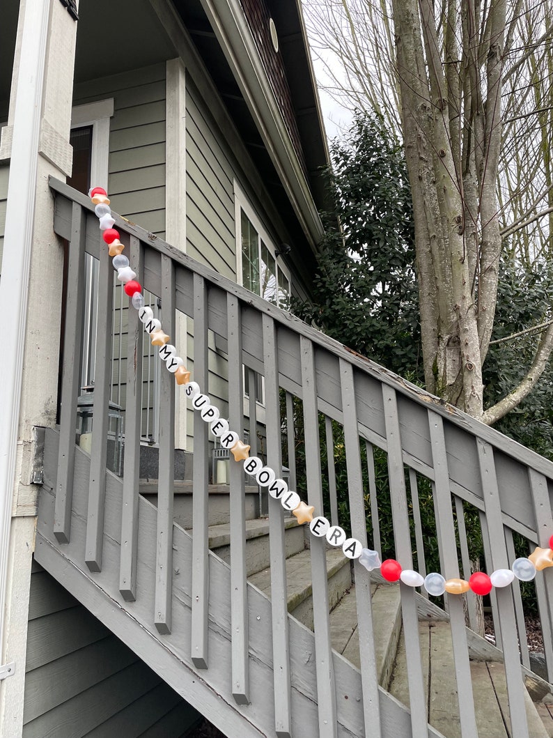 May include: A white and gold garland with the words "In My Super Bowl Era" strung on a wooden railing. The garland is decorated with red and gold balloons.
