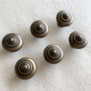 Puede incluir: Un juego de seis pomos redondos para armarios hechos de metal con tono de latón antiguo. Cada pomo presenta un diseño circular concéntrico escalonado con un punto central elevado, creando un perfil que sube desde la base hasta un remate superior.