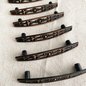 Może przedstawiać: Zestaw sześciu uchwytów do szafek w stylu antycznym. Każdy uchwyt ma zakrzywiony, prostokątny kształt z misternymi detalami w ciemnym brązie. Posiadają czarne cylindryczne słupki do montażu.