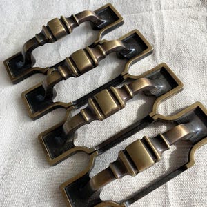 Puede incluir: Cuatro tiradores de armario antiguos de bronce con una placa trasera rectangular y un asa curvada. Los tiradores tienen un diseño detallado y ornamentado con un acabado en bronce oscuro. Las asas miden unos 10 cm de largo.