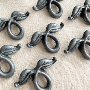 Könnte beinhalten: Nahaufnahme von mehreren grauen Metall-Serviettenringen in Blattform. Jeder Ring hat ein stilisiertes Blattdesign und eine geschwungene Basis. Die Serviettenringe sind auf einer neutralfarbenen Stoffoberfläche angeordnet.
