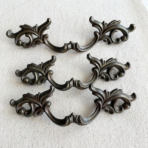 May include: Six antique bronze-coloured drawer pulls. Each pull has an ornate, symmetrical design with scrolling leaf and floral details. The pulls have a curved handle, suitable for furniture restoration or decorative purposes.