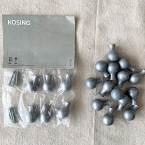 Peut inclure: Ensemble de boutons de meubles gris en forme de vase, avec des vis et un emballage portant l'inscription "KOSING". Les boutons sont disposés sur une surface de ton neutre.