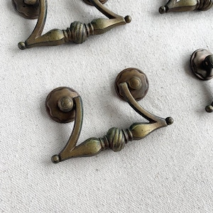 May include: Vintage brass drawer pulls with an ornate design. The pulls have a curved handle and a round backplate. The metal has a vintage, aged appearance, with colour variations from gold to bronze. Suitable for furniture restoration or adding a vintage touch to cabinets.