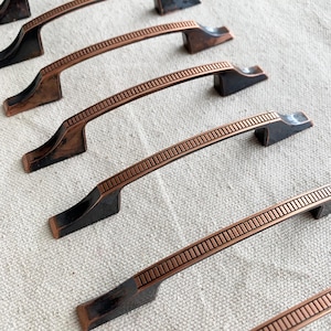 Op de afbeelding: Close-up van verschillende bronskleurige kastgrepen. De grepen hebben een rechthoekige vorm met een decoratief, getextureerd midden en donkerbronzen uiteinden. De hardware is gerangschikt op een neutraal gekleurde stof.