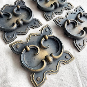 May include: Close-up of several antique bronze drawer pulls. Each pull features a decorative backplate with a curved handle. The pulls have a distressed finish, with hints of gold and dark grey. The hardware is likely for furniture restoration.