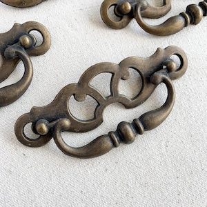May include: Close-up of several antique brass drawer pulls. Each pull features an ornate design with a curved handle and decorative backplate. The pulls have a warm, aged patina, suggesting vintage or retro style. The background is a neutral, textured surface.