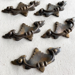 May include: Five antique bronze drawer pulls with ornate, leaf-shaped backplates and curved handles. The pulls have a dark, aged finish, suggesting vintage hardware. Suitable for furniture restoration or adding a classic touch to cabinets.