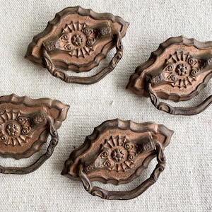 May include: Four antique bronze drawer pulls. Each pull features a decorative, scalloped backplate with a central floral design and a curved handle. The pulls have a weathered, aged appearance, with variations in color from bronze to copper.
