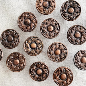 May include: Close-up of several antique bronze-colored buttons. Each button features a central raised circle surrounded by an ornate, swirling leaf and scroll design. The buttons are arranged on a neutral, textured surface.