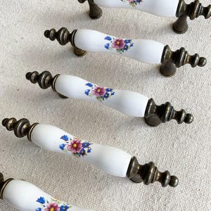 May include: A collection of white ceramic cabinet handles featuring a floral pattern in pink, purple, and blue. The handles have antique brass-coloured metal fittings. The floral design is centred on the white ceramic handle.
