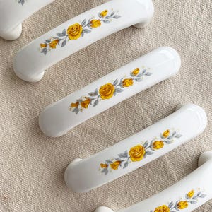 Pode incluir: Puxadores de armário de cerâmica branca com design curvo. Cada puxador é decorado com um padrão floral de rosas amarelas e folhas cinzentas. Os puxadores estão dispostos sobre uma superfície de tecido de cor neutra.