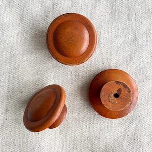 May include: Three wooden cabinet knobs with a warm, reddish-brown finish. The knobs have a circular design with a raised center and a rounded edge. The knobs are set against a neutral, textured background.