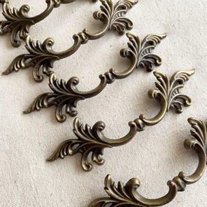 May include: Close-up of several antique bronze-colored drawer pulls. Each pull features an ornate, symmetrical design with curved handles and decorative leaf accents. The pulls are arranged on a neutral-colored surface.