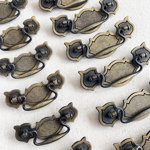 Peut inclure: Ensemble de poignées de tiroir de style antique. Chaque poignée présente une plaque arrière décorative en forme de bouclier avec une poignée incurvée. Le métal a une finition bronze avec des accents sombres, suggérant un aspect vieilli. Les poignées sont disposées sur une surface claire.